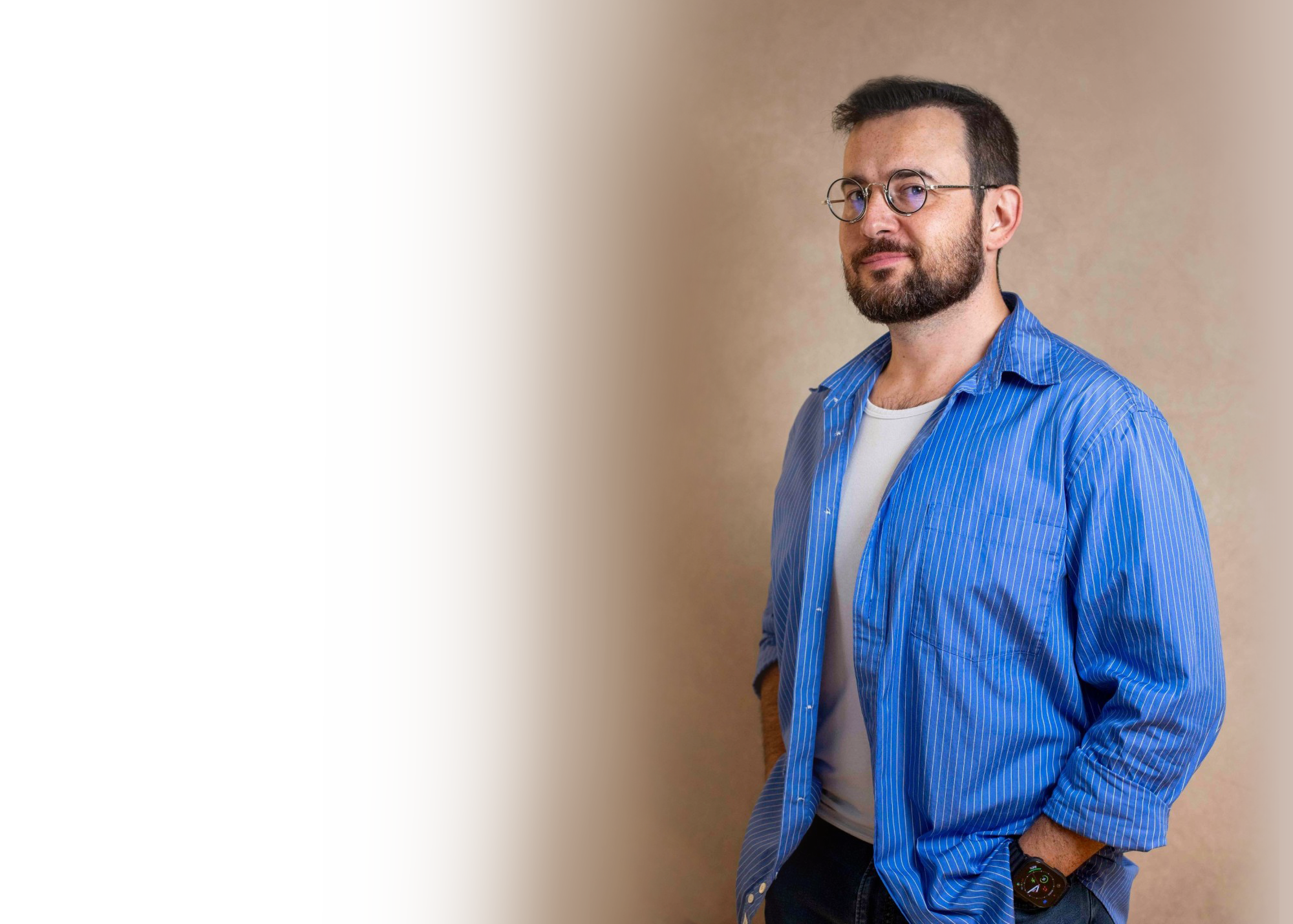 A man with dark hair and a beard wearing glasses, a blue striped shirt, and a white t-shirt, standing with his hands in his pockets against a beige background.