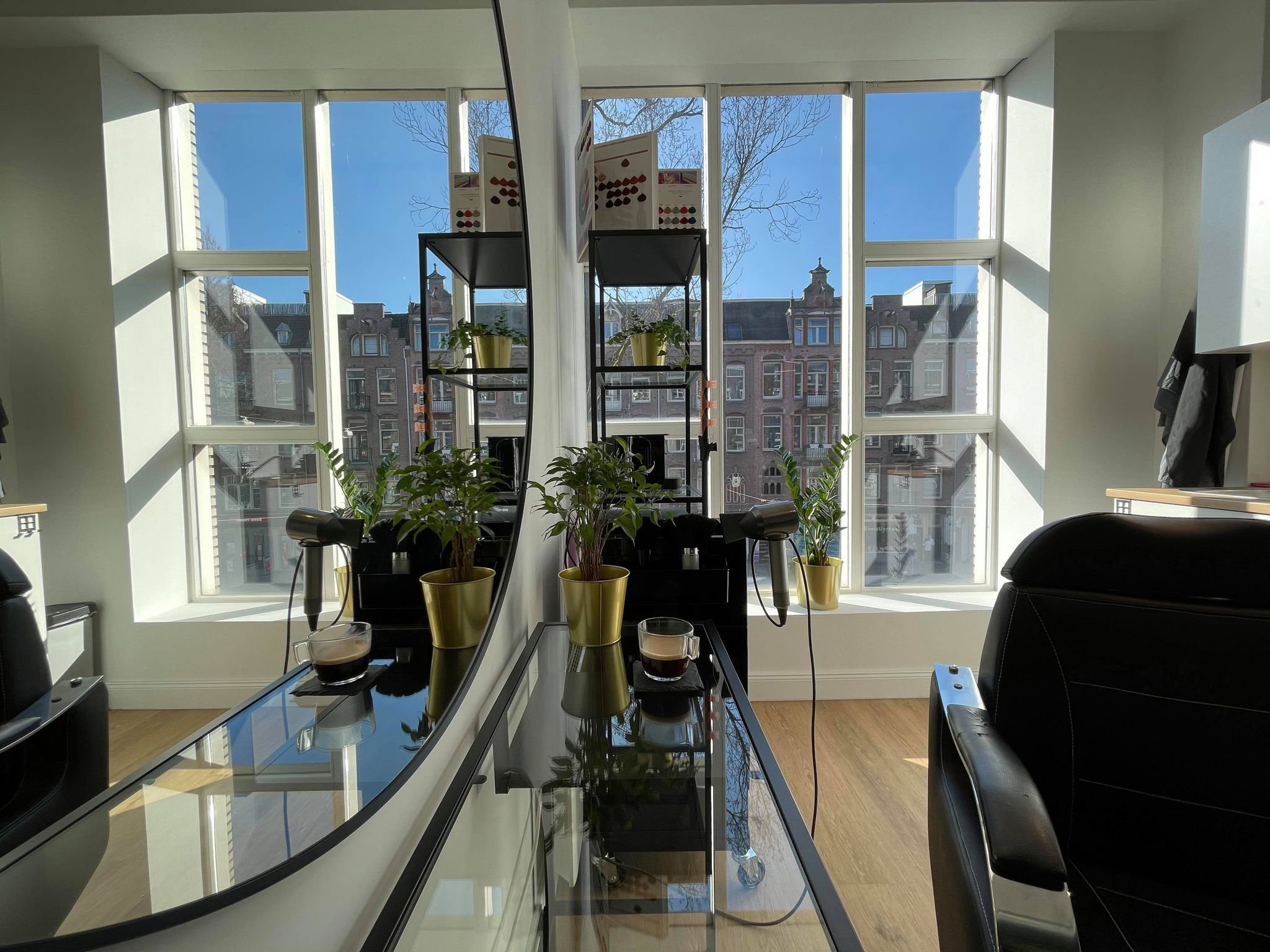 Interior of an office with large windows, green plants in gold pots, a mirror, and a glass table with a coffee cup, hairdryer, and other items, overlooking a cityscape with buildings and blue sky.