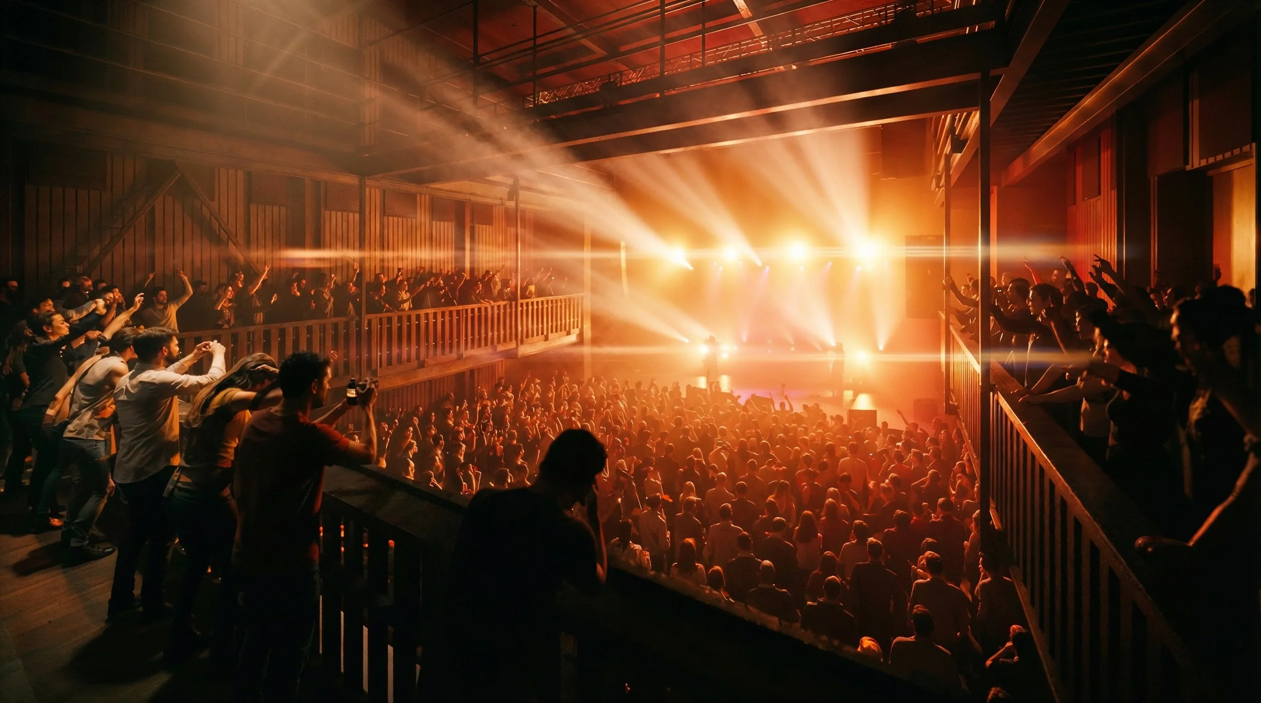 Foule dans une salle de concert ou de spectacle avec des lumières orange brillante à l'arrière, la scène est remplie de spectateurs. Des personnes regardent depuis une mezzanine sur les côtés, certaines dansent ou prennent des photos.