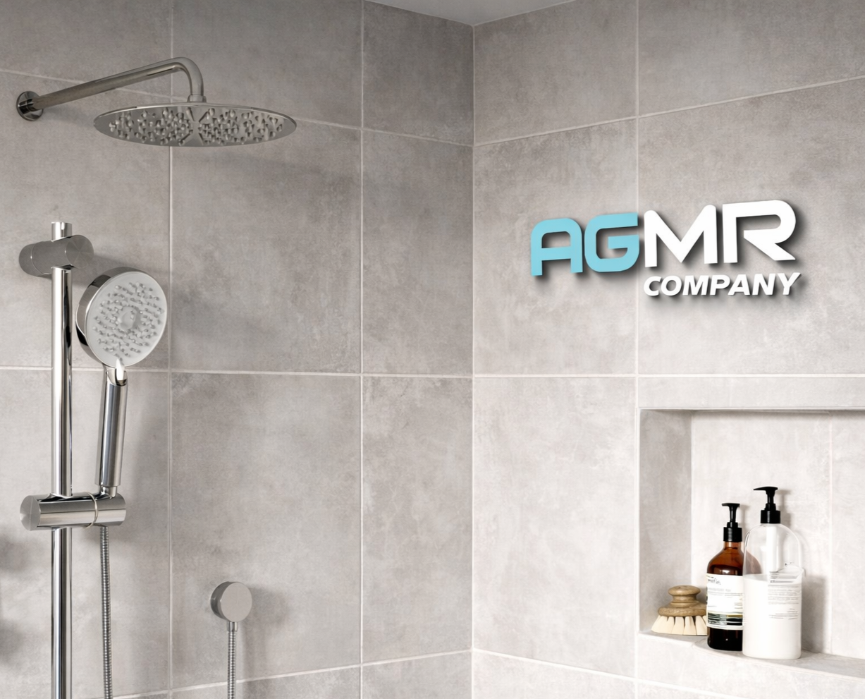 A modern shower area with a gray tiled wall, a rainfall showerhead, a handheld showerhead, and a built-in shelf holding two bottles of soap or shampoo and a scrub brush. The image also features the text 'AGMR COMPANY' in blue and white font on the wall.