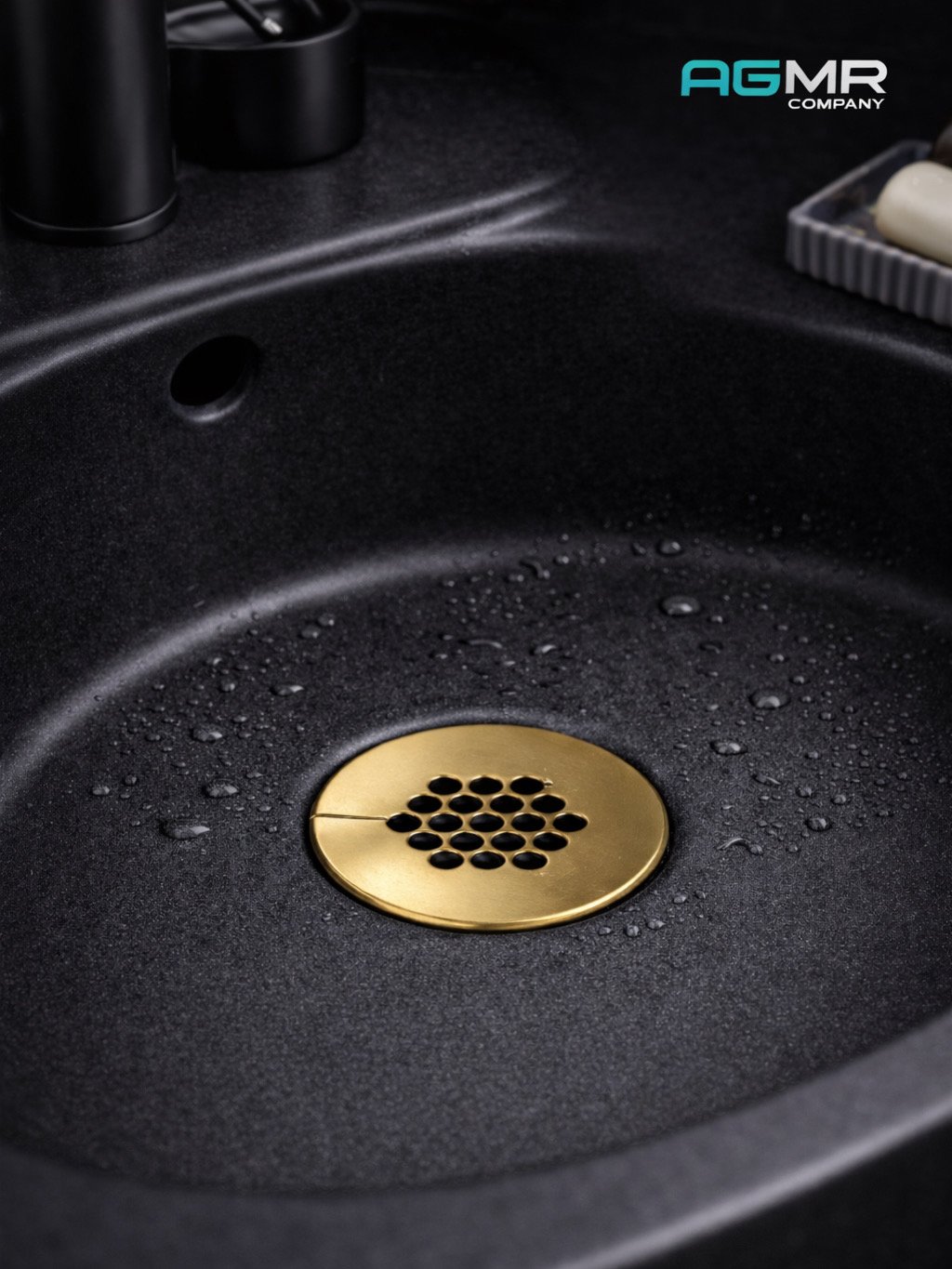 Close-up of a black kitchen sink with droplets of water and a gold drain cover, with a soap dish and bottles nearby.