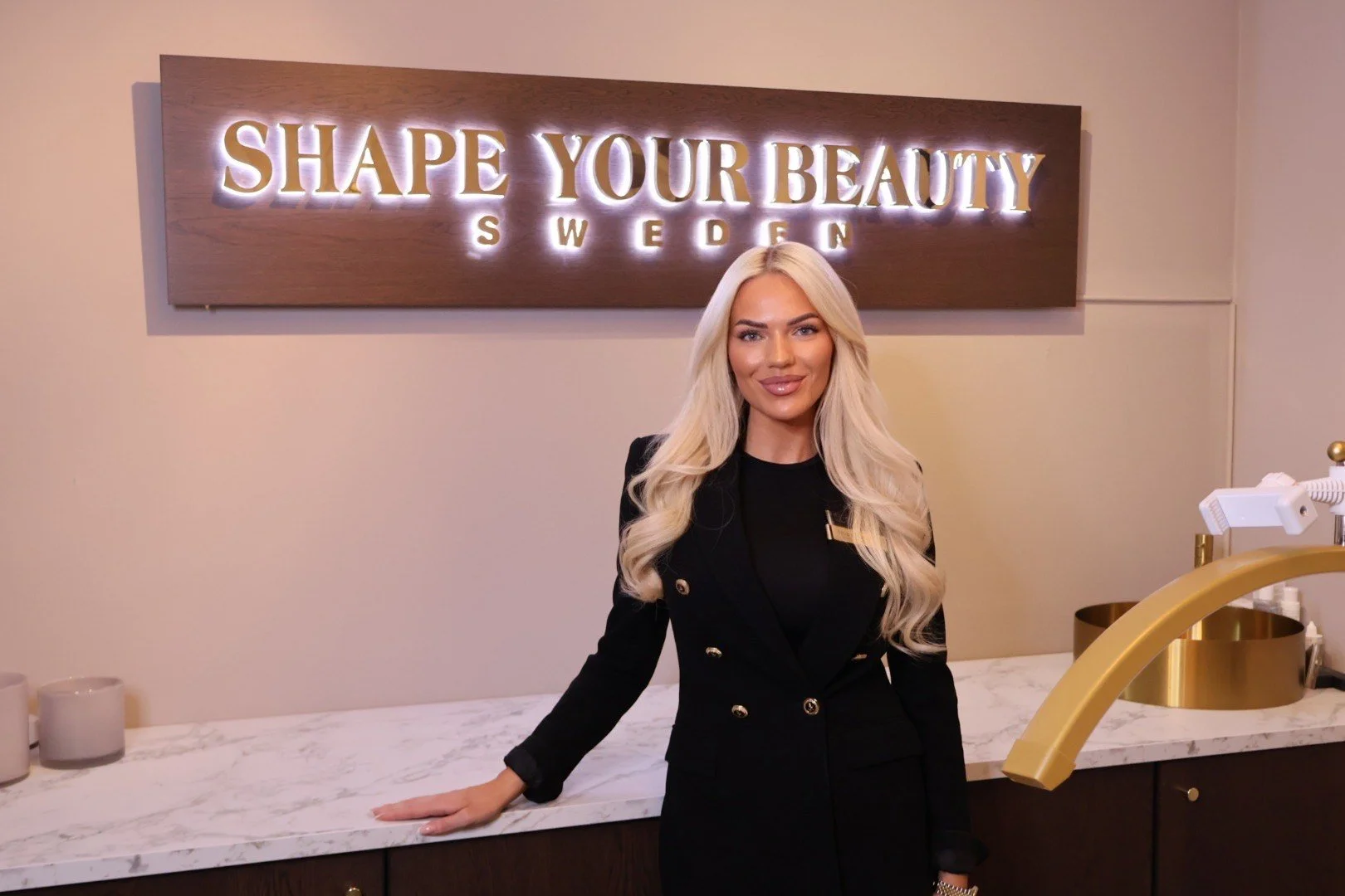 En kvinna med långt blont hår står framför en vägg med en ljusskylt som säger 'SHAPE YOUR BEAUTY SWEDEN'.