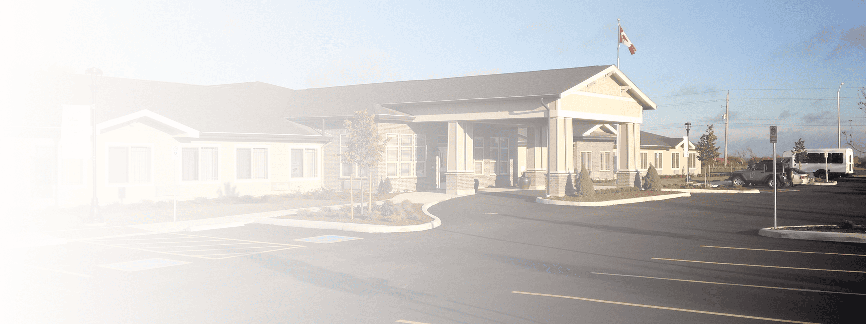 A large, modern building with a tower in the front, surrounded by a parking lot with several cars, a flagpole with a Canadian flag, and a few small trees.