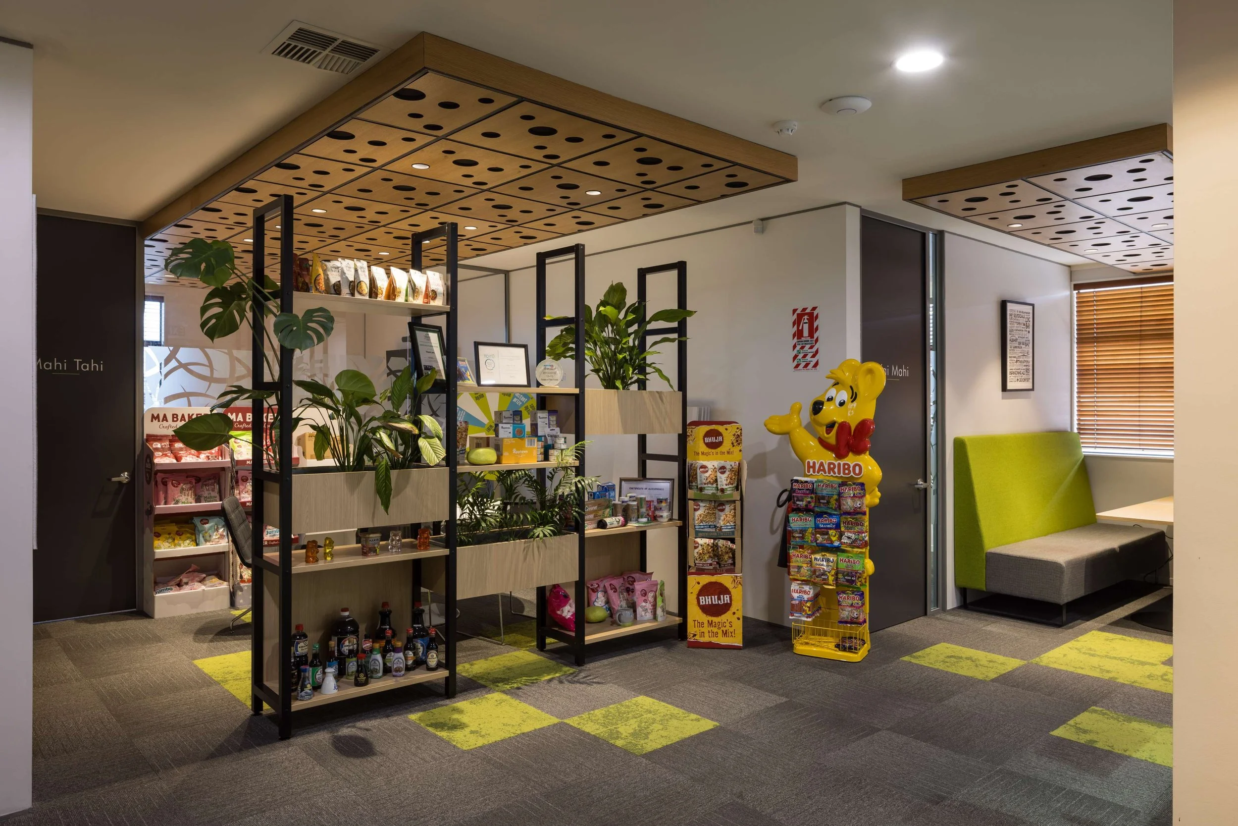 Indoor area with shelves displaying snacks and displays, a yellow Haribo mascot statue, a green and gray booth seating area, and various plants.