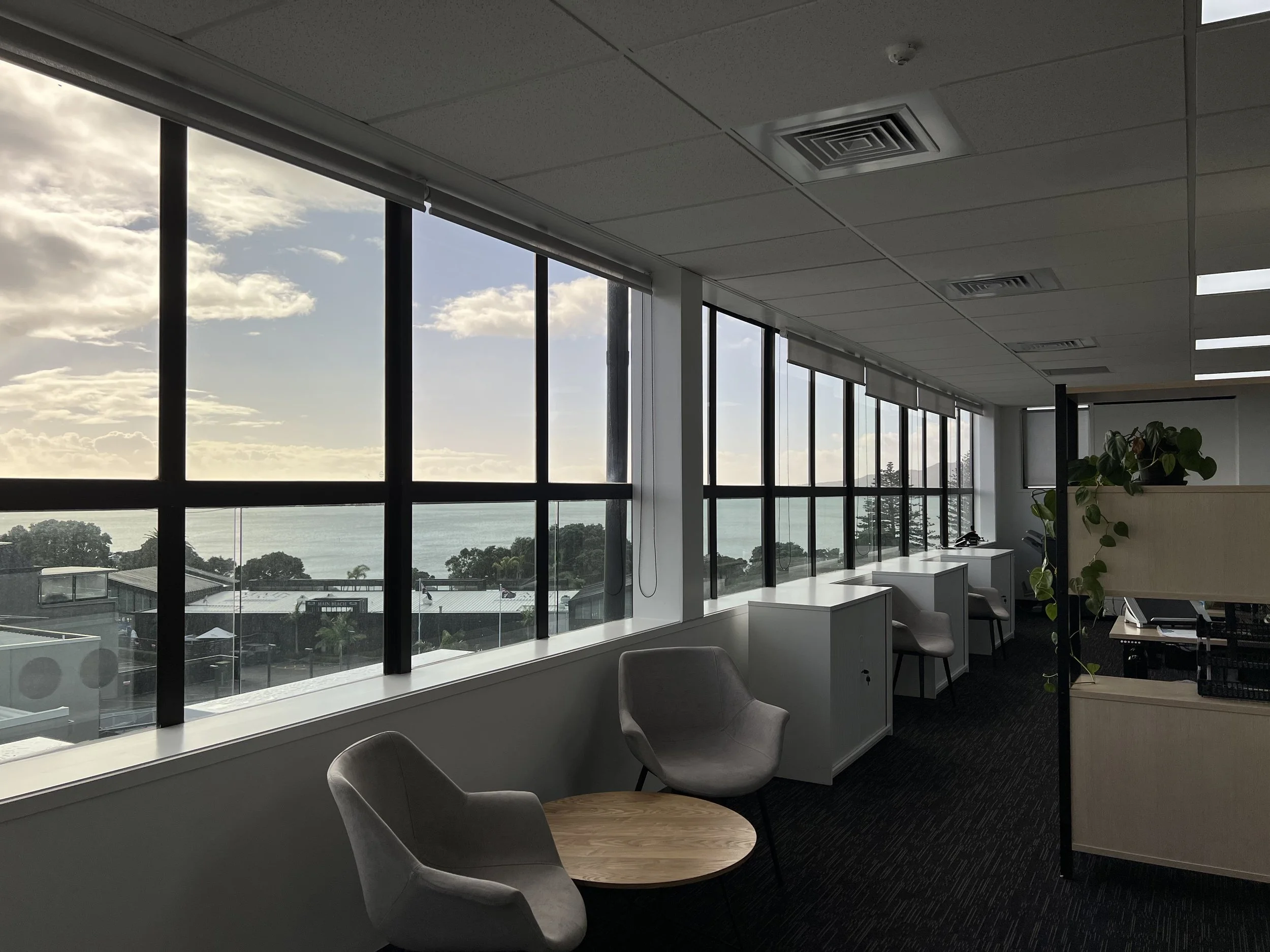 Empty modern office with large windows showing a view of the sky, water, and trees, furnished with chairs, small desks, and potted plants.