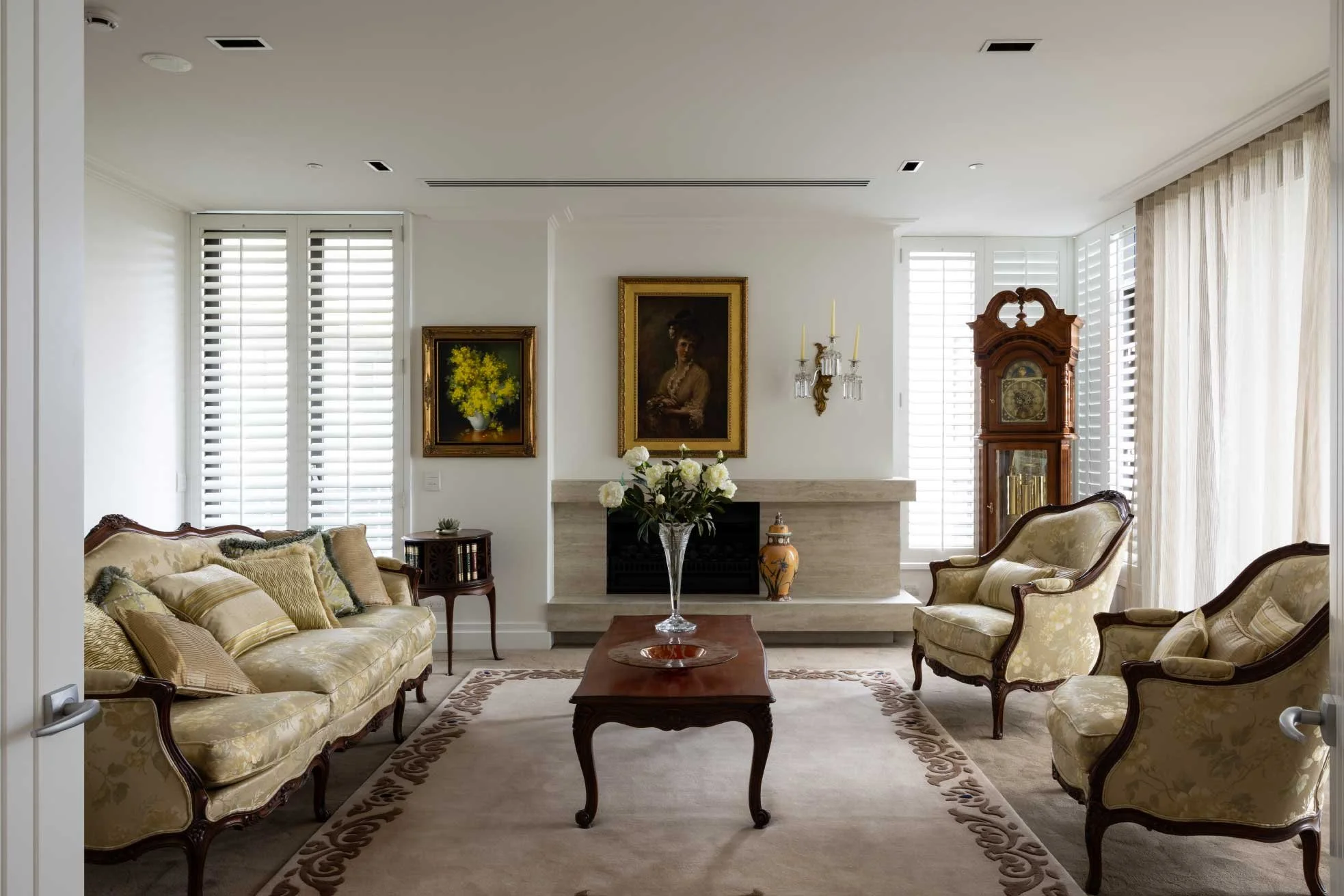 Elegant living room with vintage style furniture, a central coffee table with a vase of white flowers, a stone fireplace, wall paintings, and tall windows with white shutters and curtains.
