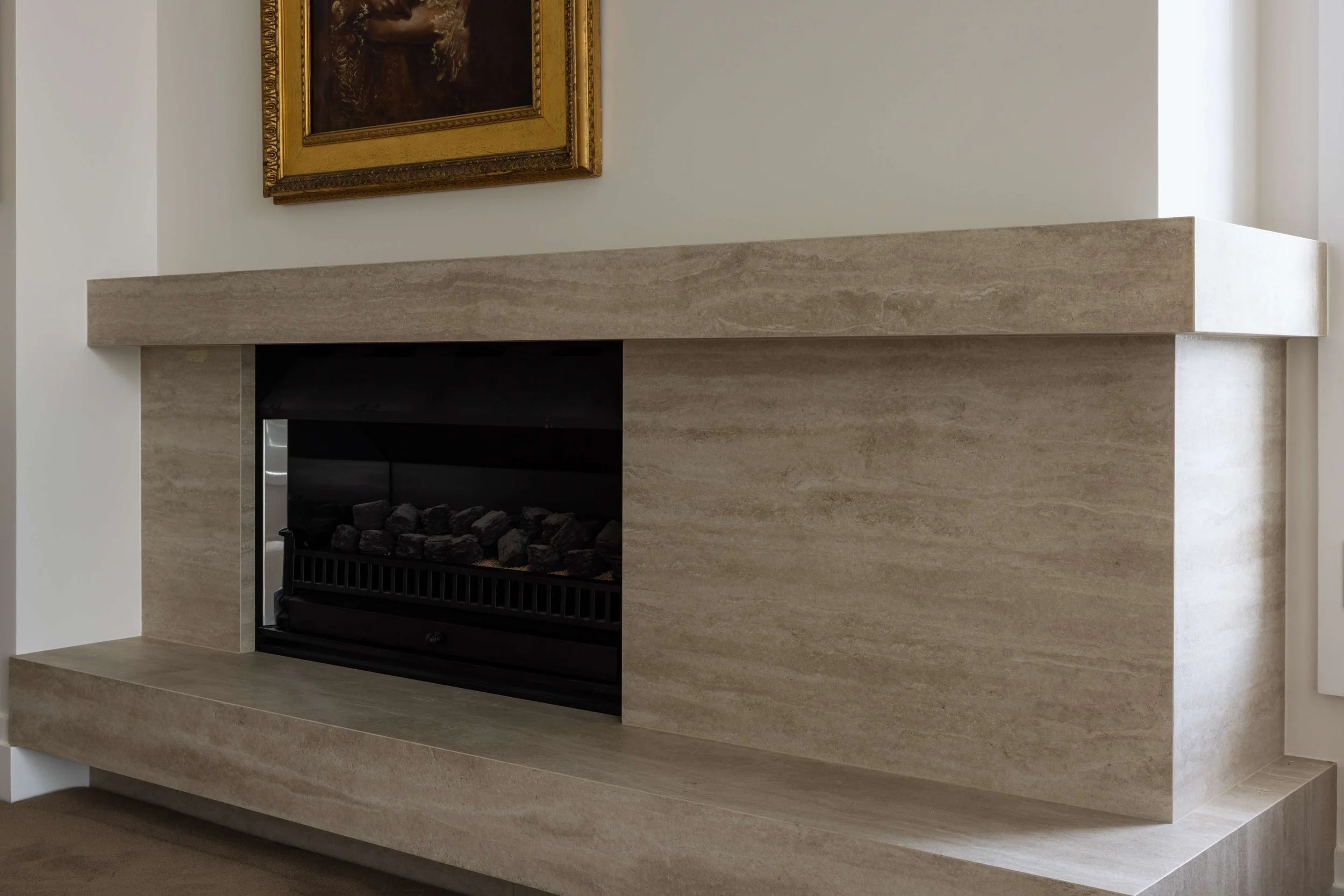 Modern fireplace with beige stone surround and a black interior, above a white wall with a framed painting or picture.