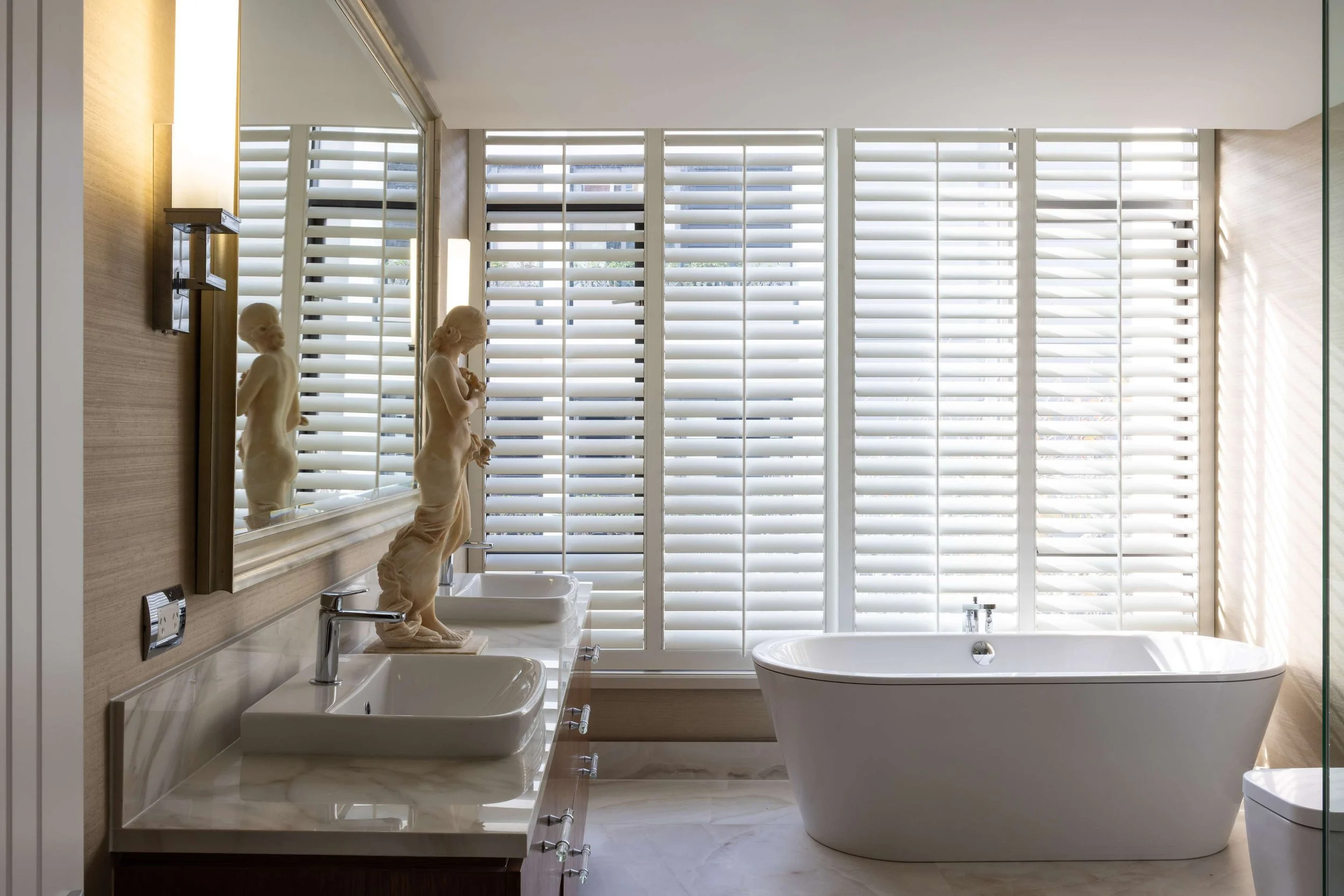 Modern bathroom with a large bathtub, two sinks with decorative statues, a mirror, and window shutters.