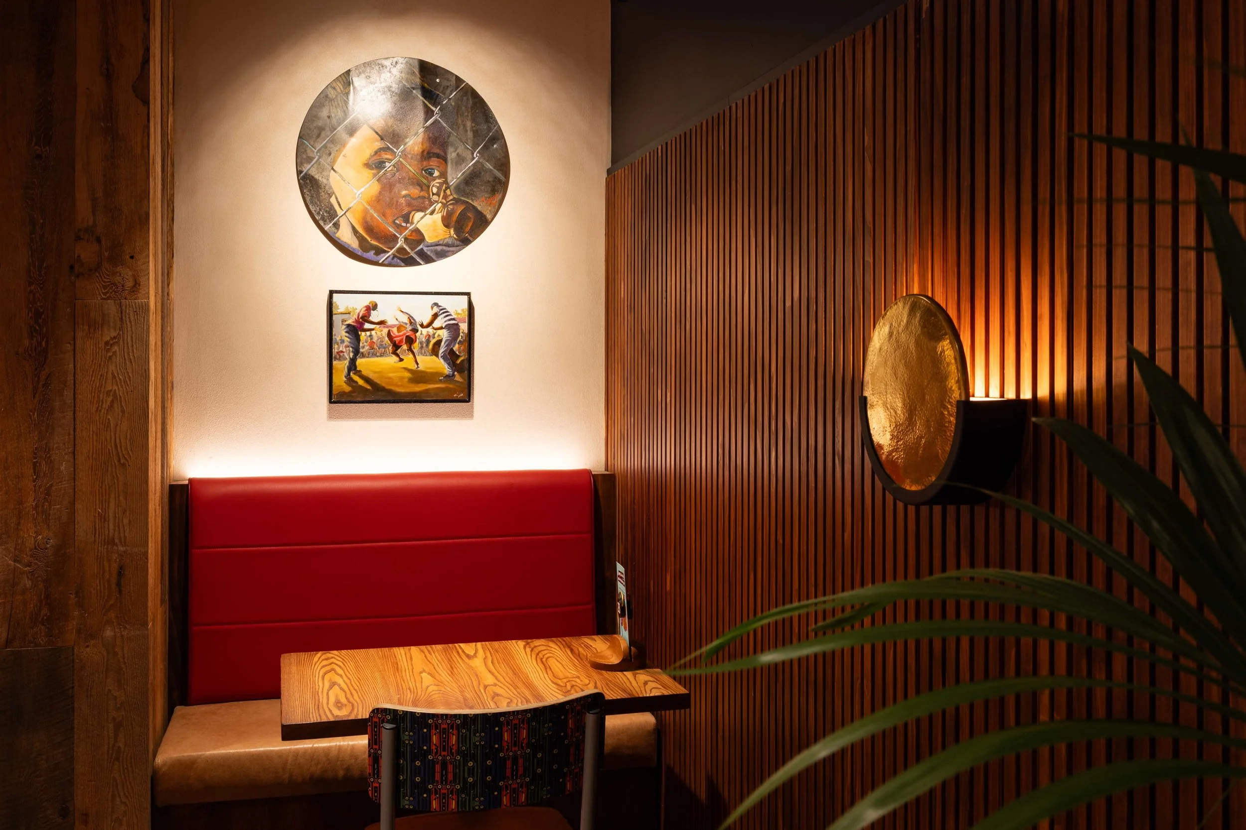 A cozy dining corner with a brown wooden table and a red cushioned booth. The wall features artwork including a large circular painting of a woman speaking into a microphone and a smaller rectangle painting of people dancing. The right side has a textured wooden wall with a gold semi-circular wall sconce emitting warm light. Green plant leaves are visible in the foreground.