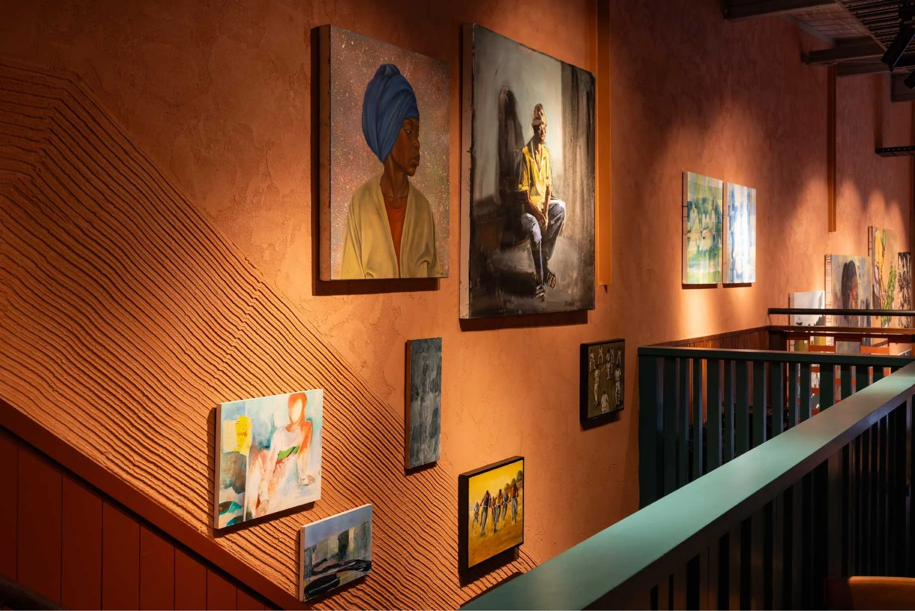 Interior view of an art gallery with various paintings displayed on an orange textured wall, including portraits and landscapes.