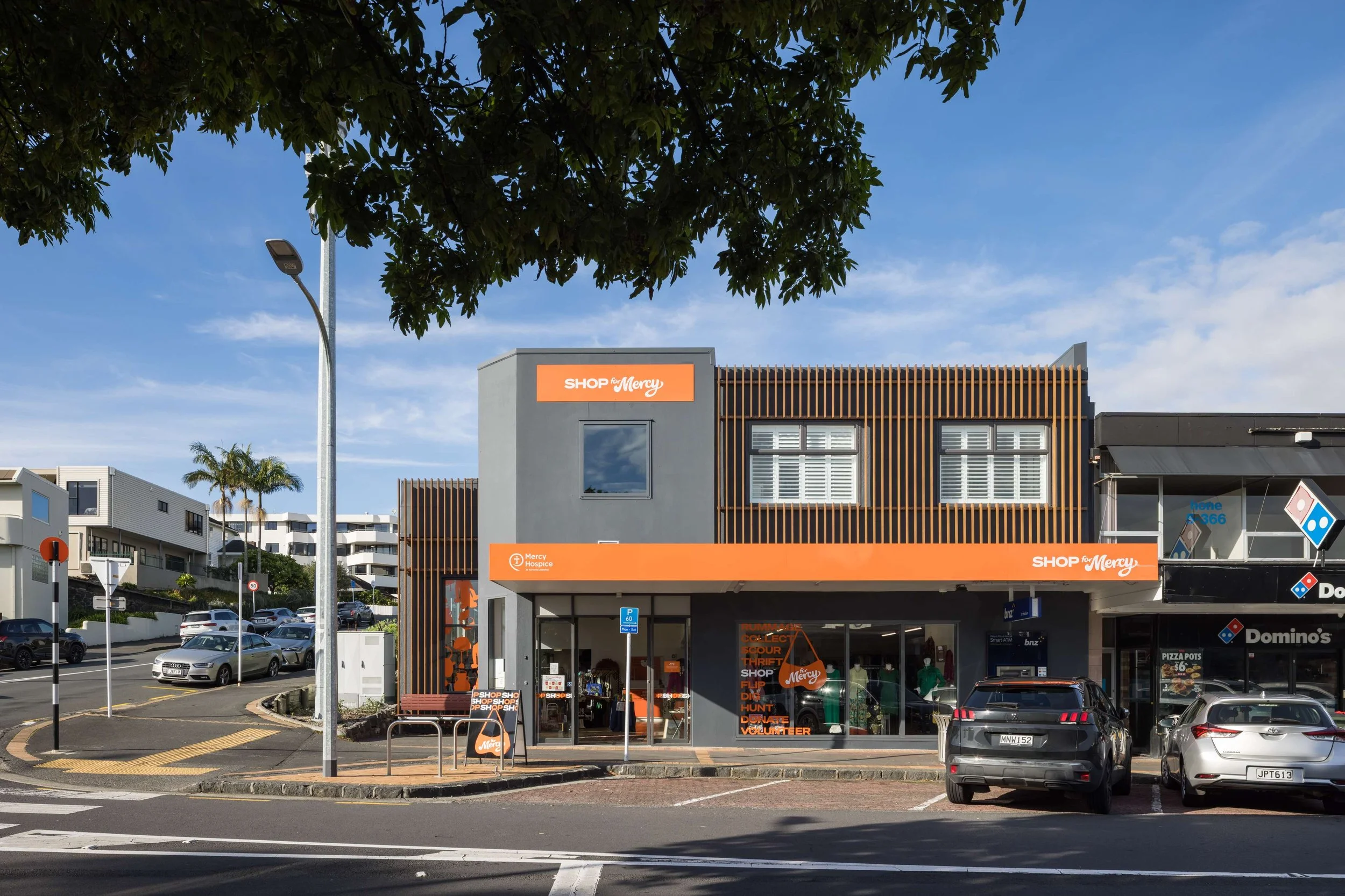 A two-story commercial building with orange signs reading 'Shop for Mercy' and 'Mercy Hospice.' The ground level has a glass storefront with various items inside, and there are parked cars in the parking lot in front. The street is lined with trees and a street light, under a partly cloudy sky.