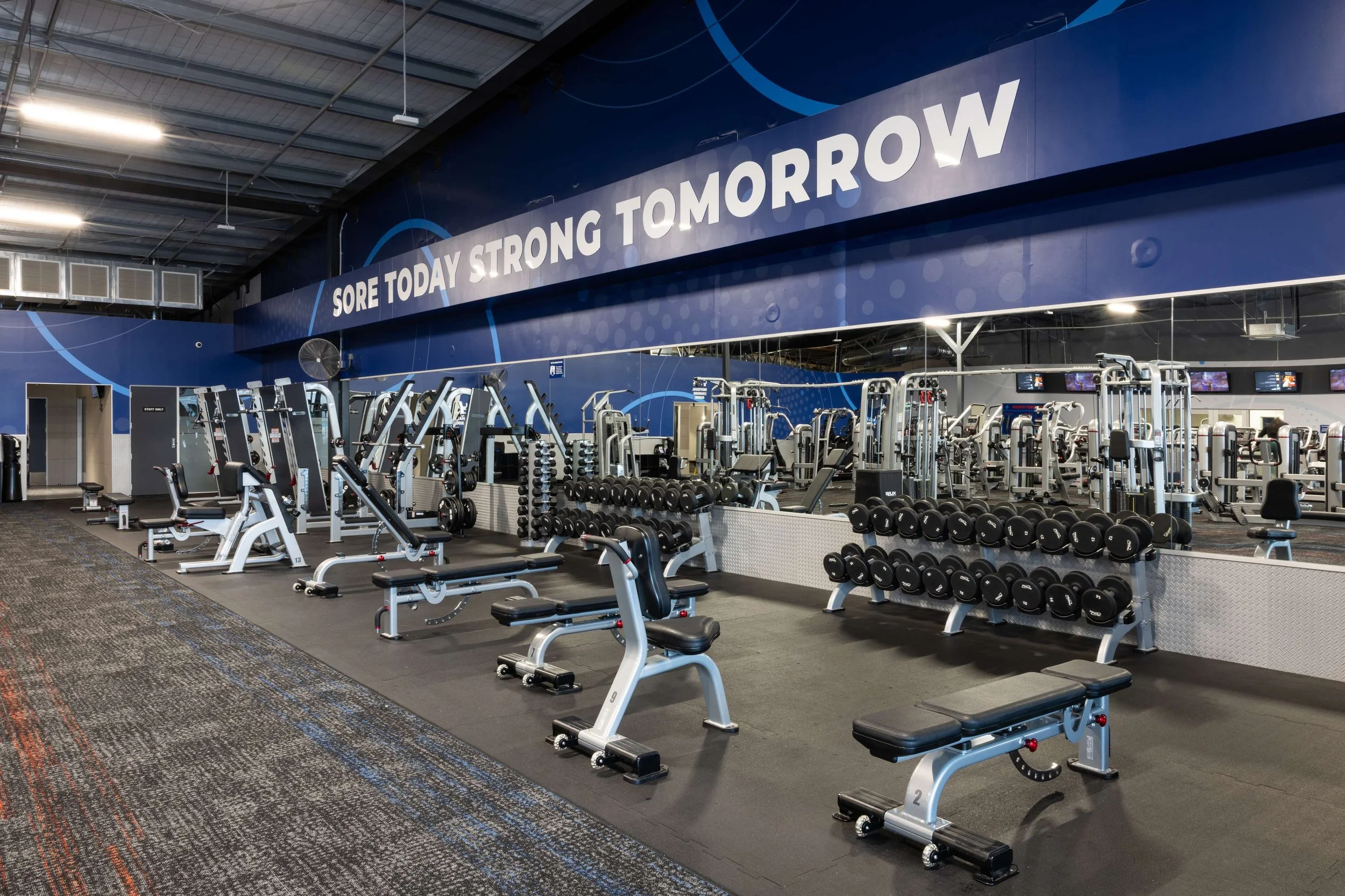 Empty gym with various workout equipment, dumbbells, and benches, with a large blue wall featuring the motivational phrase "Sore Today Strong Tomorrow."