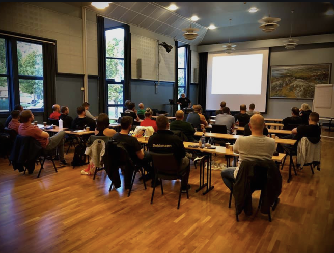 Mann som holder presentasjon foran en gruppe mennesker i et konferanserom med store vinduer, trebord og en projektor.