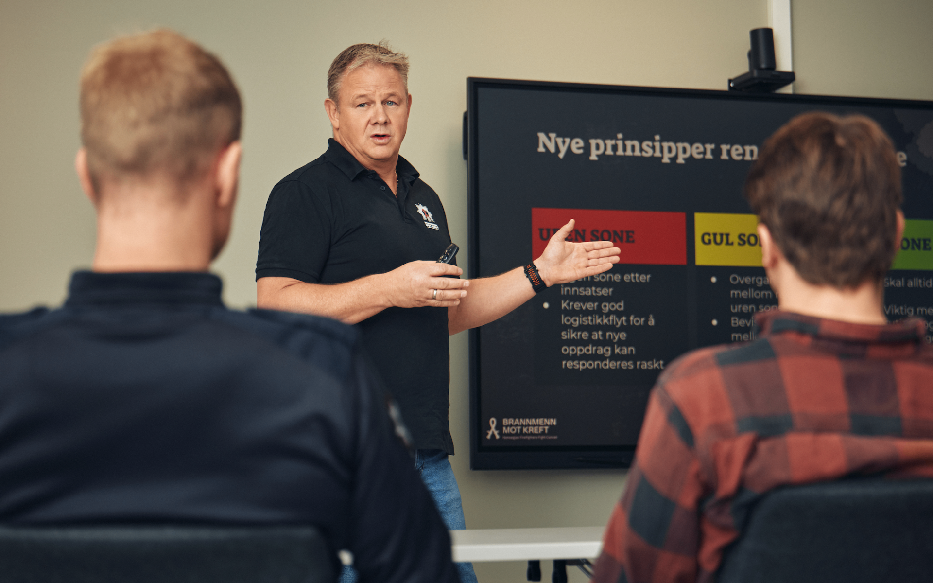 En mann som holder en fjernkontroll og peker mot en presentasjon på en skjerm foran tre personer med ryggen til, i et klasserom eller møteområde.