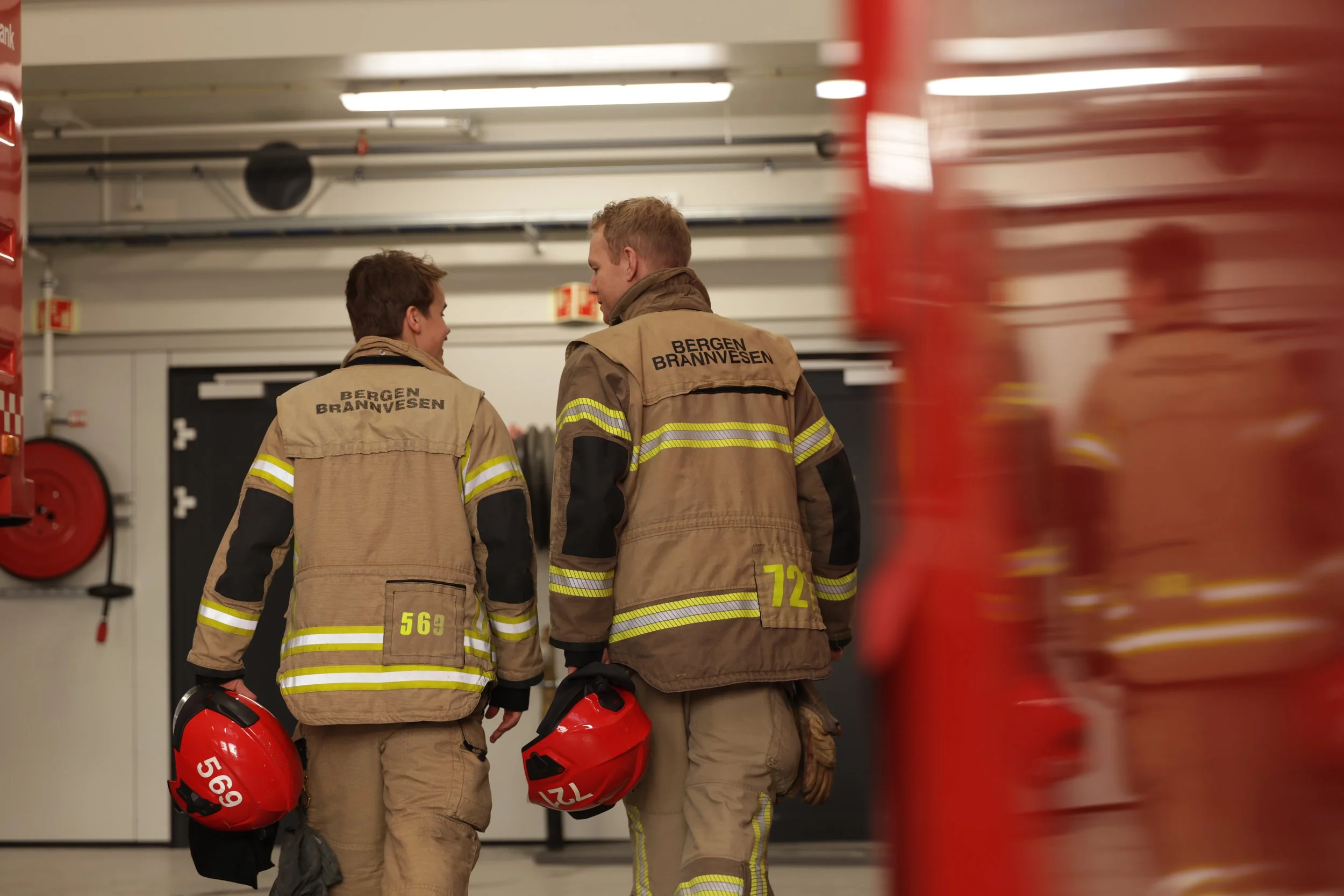 Tre brannmann i beige uniform med reflekterende striper, står i en hall med røde brannhjelmer under armen, og snakker sammen, en av dem har et hjelm med nummer 569