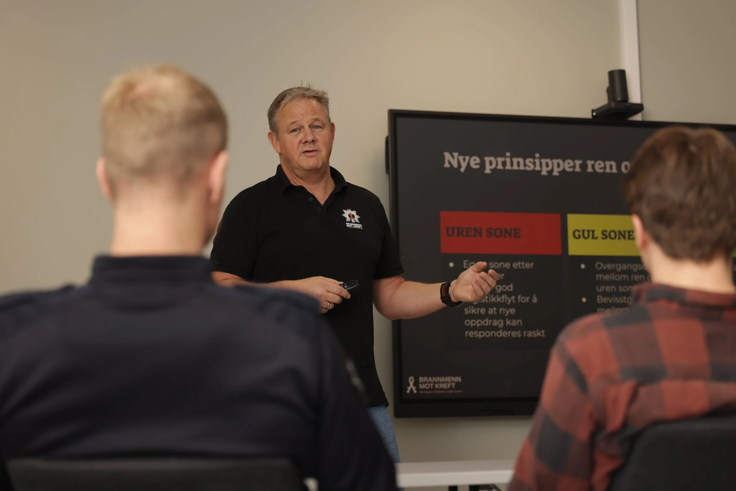 En mann holder en presentasjon foran en gruppe mennesker i et klasserom, med en plakat som viser prinsipper for ren og urenset jord. Flere personer er synlige fra ryggen, og det er en stor skjerm med tekst i bakgrunnen.