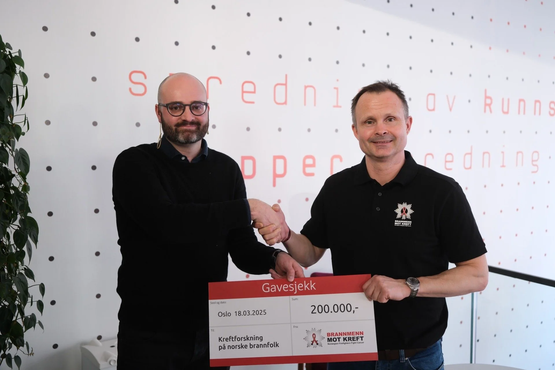 To menn står foran en vegg med prikket design og tekst. Den ene mannen holder et gavesjekk på 200 000 kroner. De ser glade ut og håndhilser.