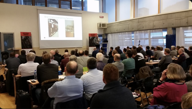 En stor gruppe mennesker deltar i et møte eller konferanse, der en person snakker foran en skjerm som viser en presentasjon.