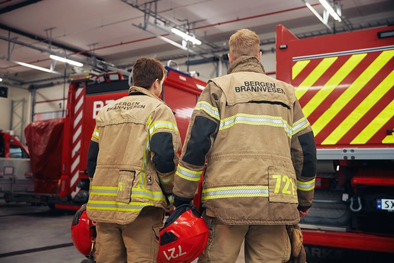 To brannmenn står med ryggen til, iført brannmannsklær, inne i en brannstasjon med brannbil i bakgrunnen.