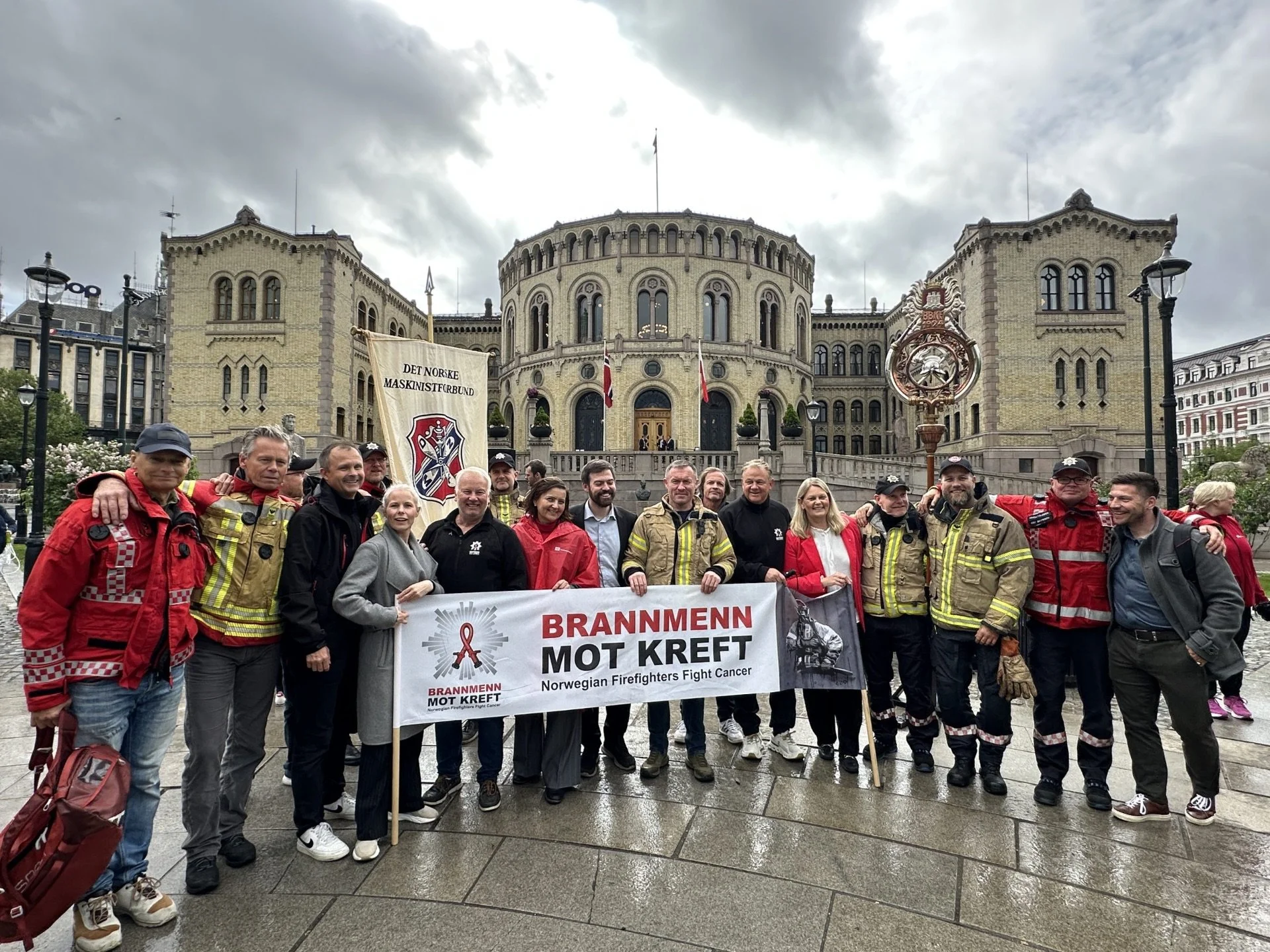Gruppemedlemmer, inkludert brandmenn, står foran Stortinget med bannere som støtter kreftforskning og mot brann. Været er overskyet og de er utendørs i Oslo.