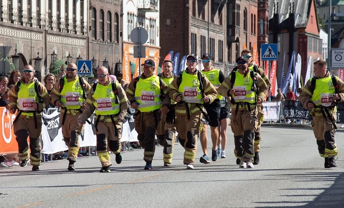 En gruppe mennesker kledd i uniformer og løpende under et arrangement på en bygate, med folk som ser på fra sidelinjen.