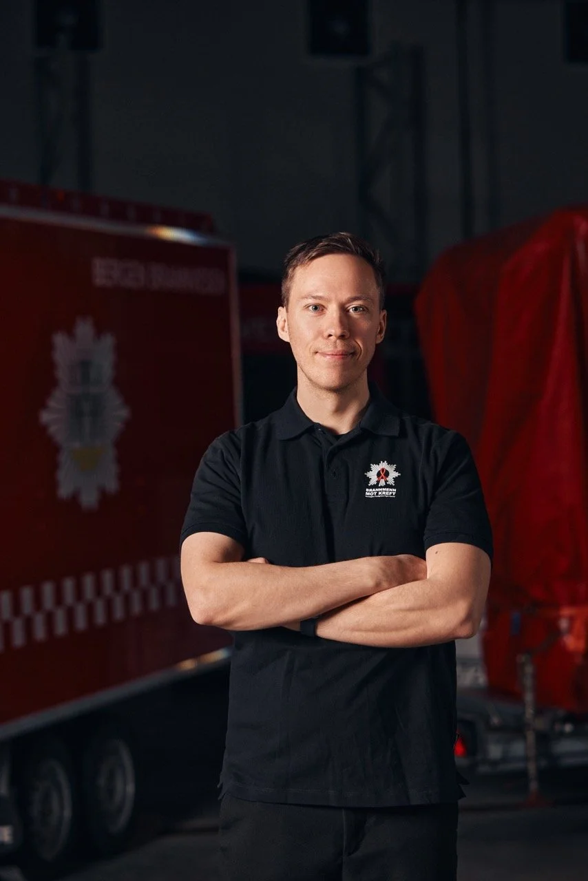 En mann med kort hår, kledd i svart polo med logo for Røde Kors, står med armene korslagt foran en rød ambulanseslukker.