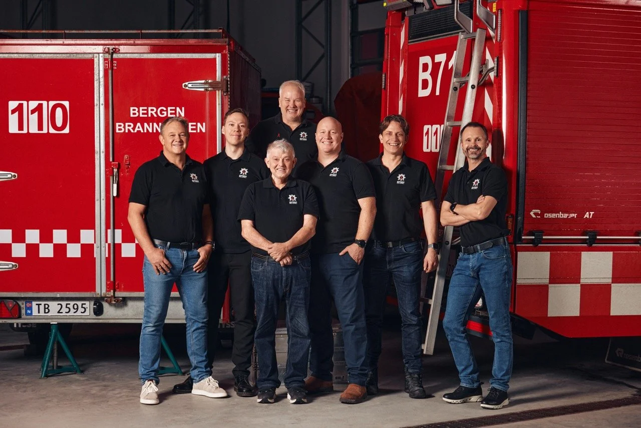 Syv personer som står foran to røde brannbil med logoer og tekst fra Bergen brannvesen, inne i et brannstudio.