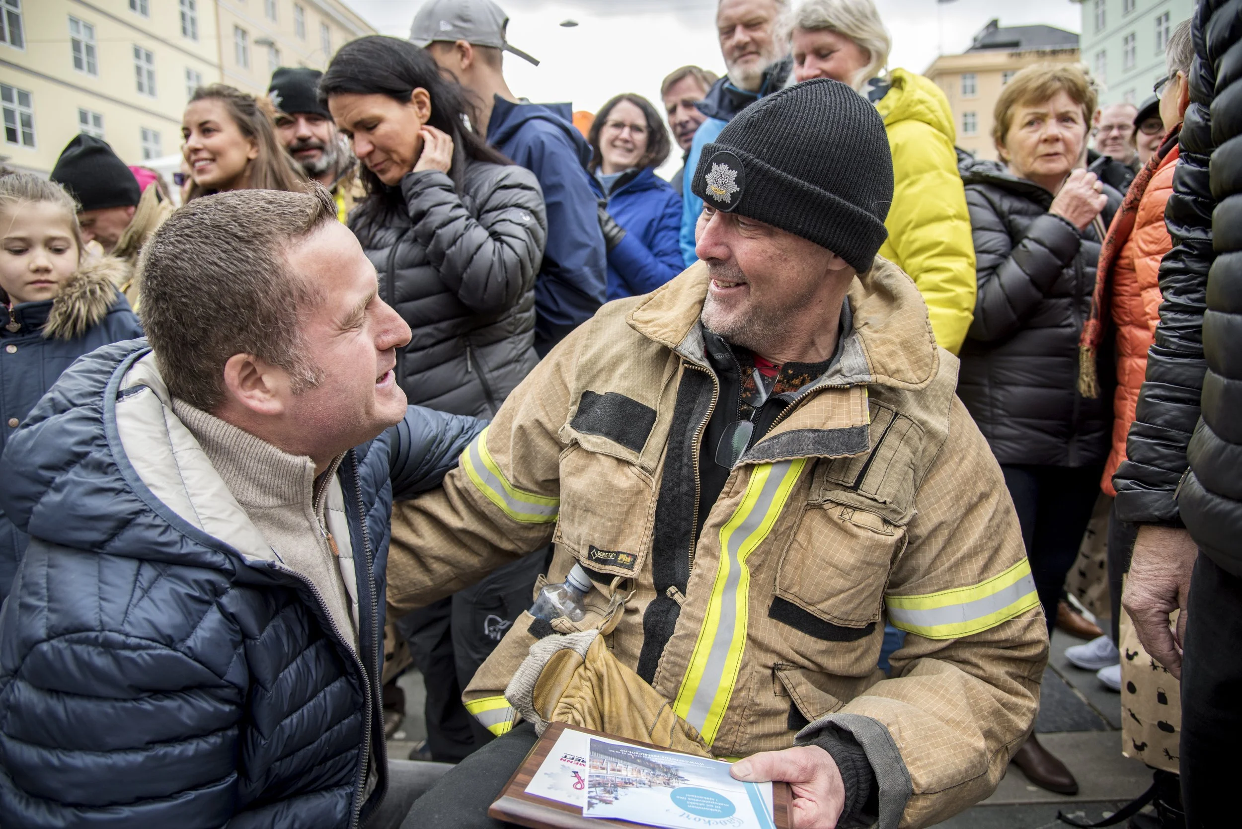 En gruppe mennesker, blant dem en mann i verneutstyr, har en samtale under en utendørs hendelse i bymiljø. Flere mennesker smiler og ser engasjerte ut.