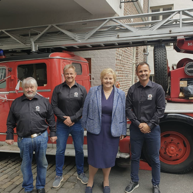 Firemenn og en kvinne står foran en rød brannbil, alle smiler, i en gate med brostein.
