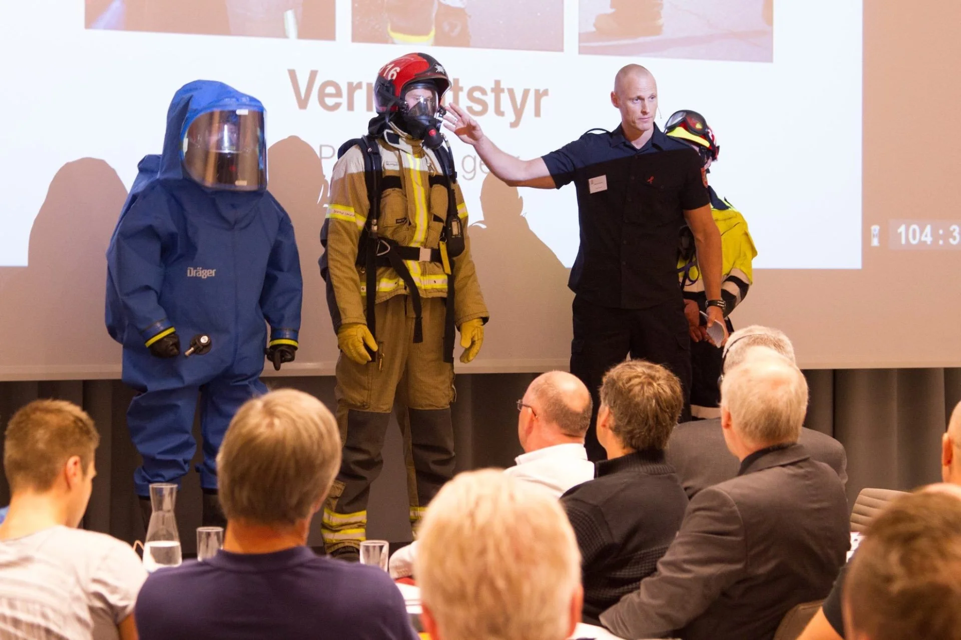 En presentasjon med to personer i spesialutstyr for brannvesen og en person som snakker til et publikum i en konferansesal. Bakgrunn med en projektor som viser bilder og tekst.