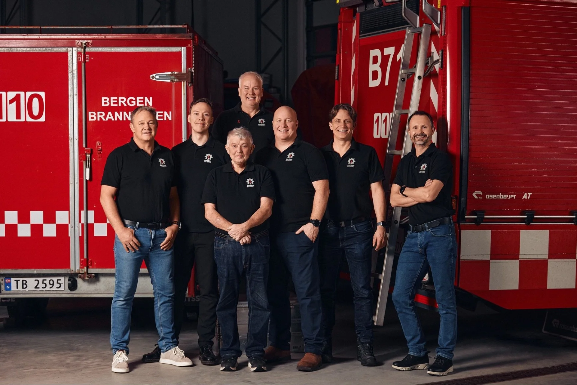 En gruppe på sju personer står foran røde brannbil, alle smiler og har på seg sorte t-skjorter med en logo for Bergen brannvesen. De står inne i et lager eller garasjeområde.