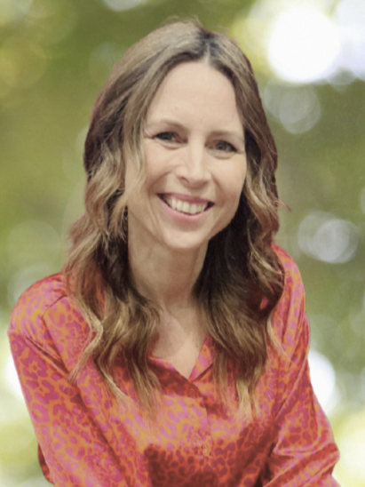 A woman with long wavy brown hair smiling outdoors in front of a blurred green background, wearing a pink and orange patterned blouse.