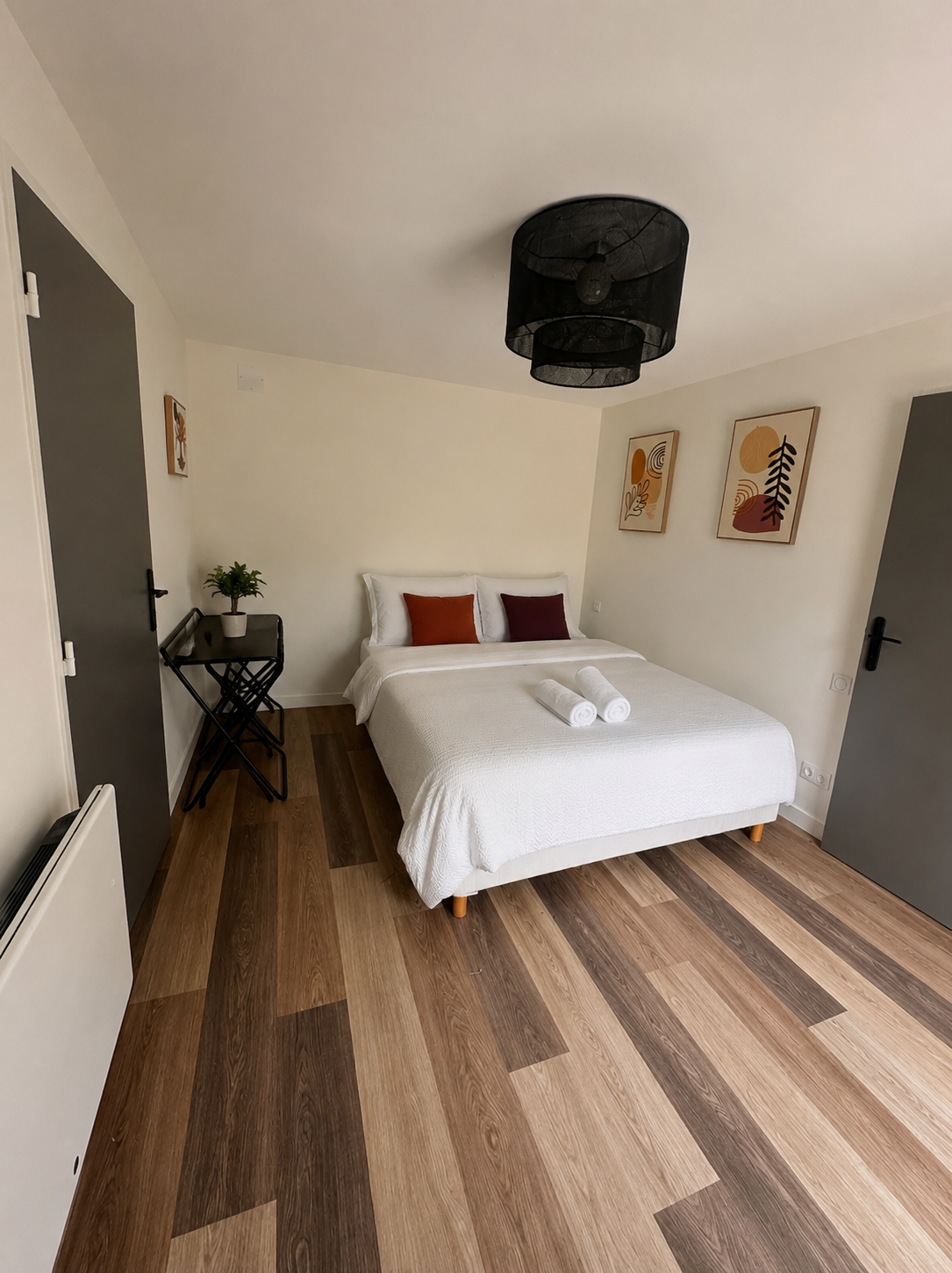 Un couloir intérieur avec un sol en bois, des murs beige avec trois tableaux représentant des paysages, et des appliques murales éclairant doucement. Au fond, une pièce avec un lit visible.