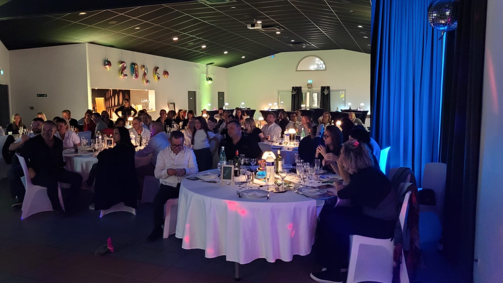 Une fête ou une réception avec de nombreuses personnes assises autour de tables rondes dans une salle décorée pour un événement de fin d'année ou une célébration, avec des décorations indiquant l'année 2026.