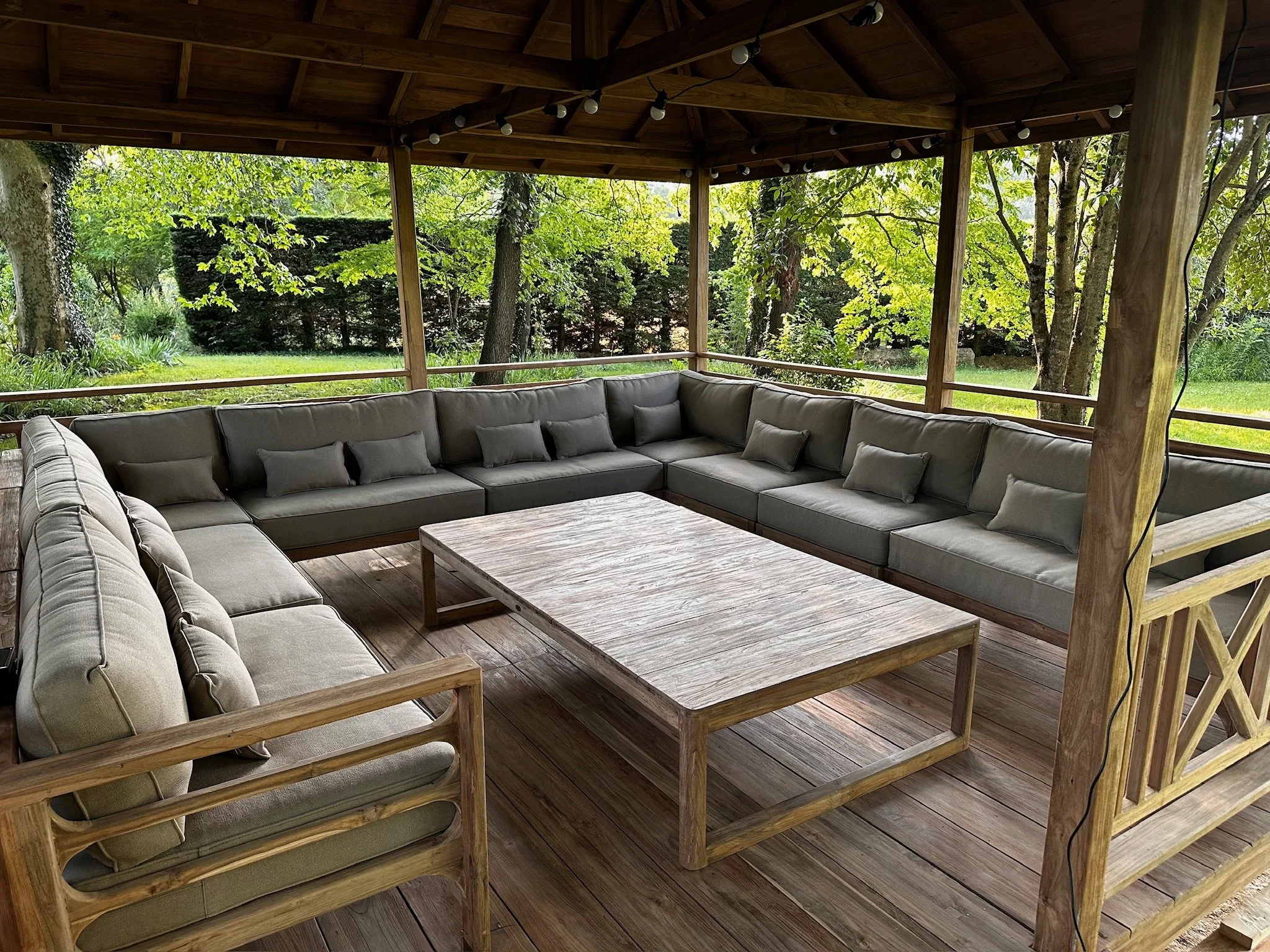 Salon de patio en bois avec canapé en canapé gris, table en bois, dans un espace ouvert entouré d'arbres verts.