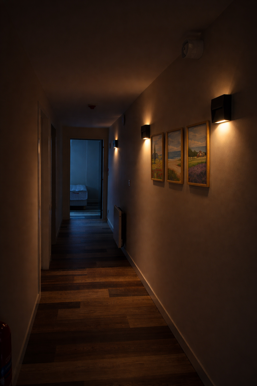 Un couloir intérieur avec un sol en bois, des murs beige avec trois tableaux représentant des paysages, et des appliques murales éclairant doucement. Au fond, une pièce avec un lit visible.