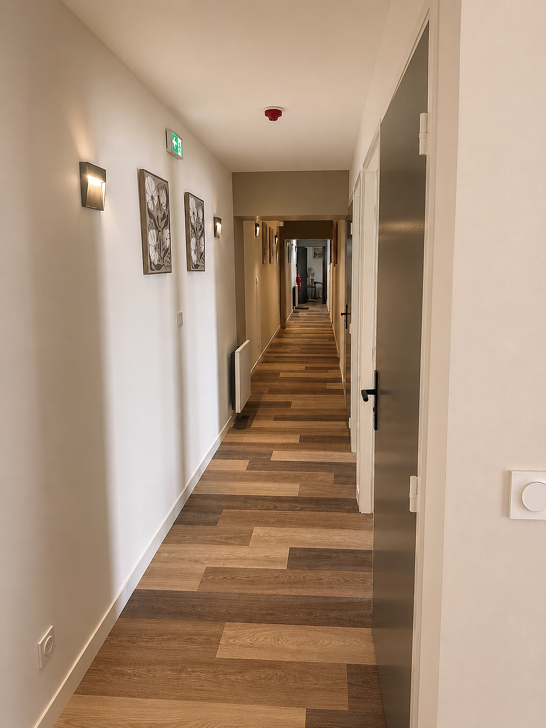 Couloir intérieur avec deux tableaux modernes, lampes murales, sol en parquet, sortie de secours et plafond avec détecteur de fumée.