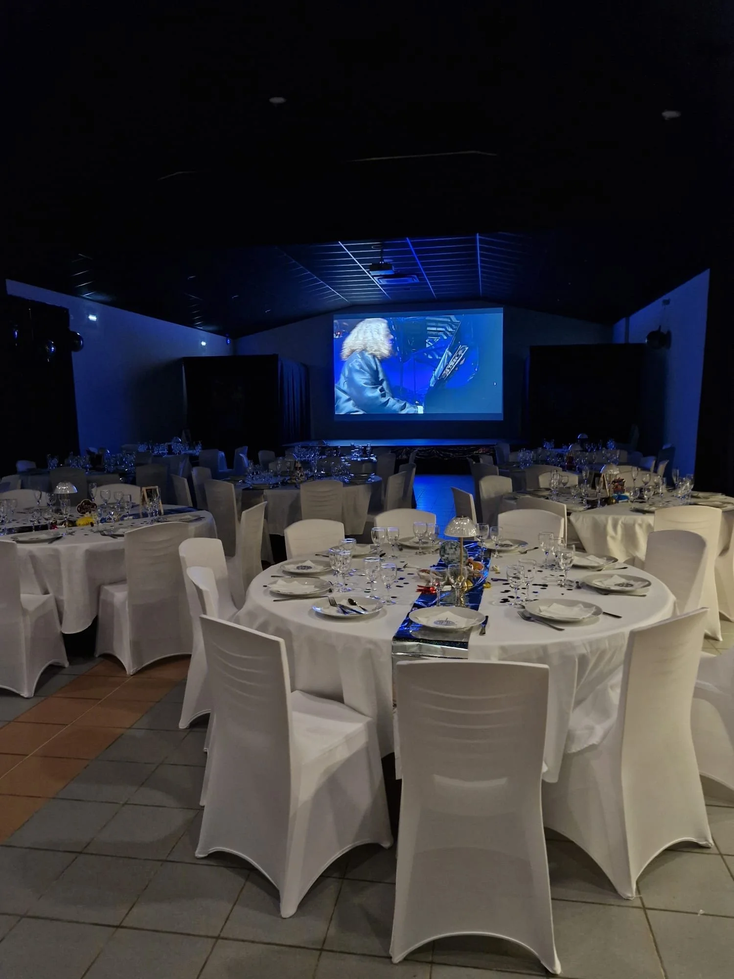 Salle de réception élégante avec tables rondes, nappes blanches, vaisselle et décorations, et un grand écran de projection.