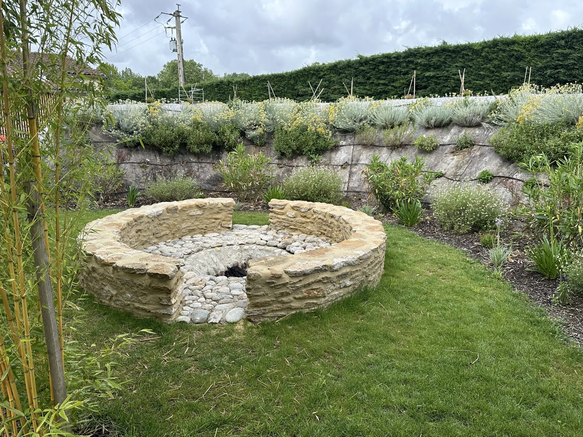 Un jardin avec un cercle en pierre, des arbustes et une haie de fleurs colorées, sous un ciel nuageux