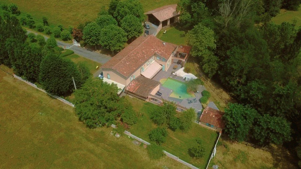Une maison entourée d'arbres, avec une piscine et une zone de détente dans le jardin, vue aérienne.
