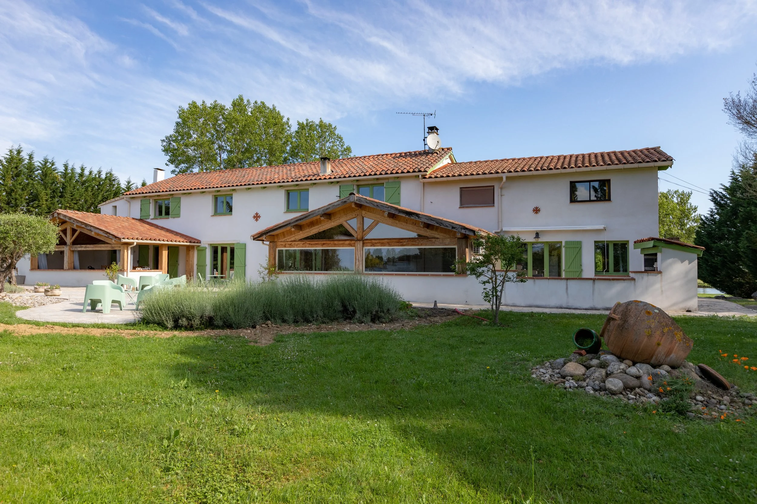 Maison de style provençal avec murs blancs, toiture en tuiles rouges, volets verts, et un jardin avec pelouse, plantes, pierres décoratives, et mobilier vert.