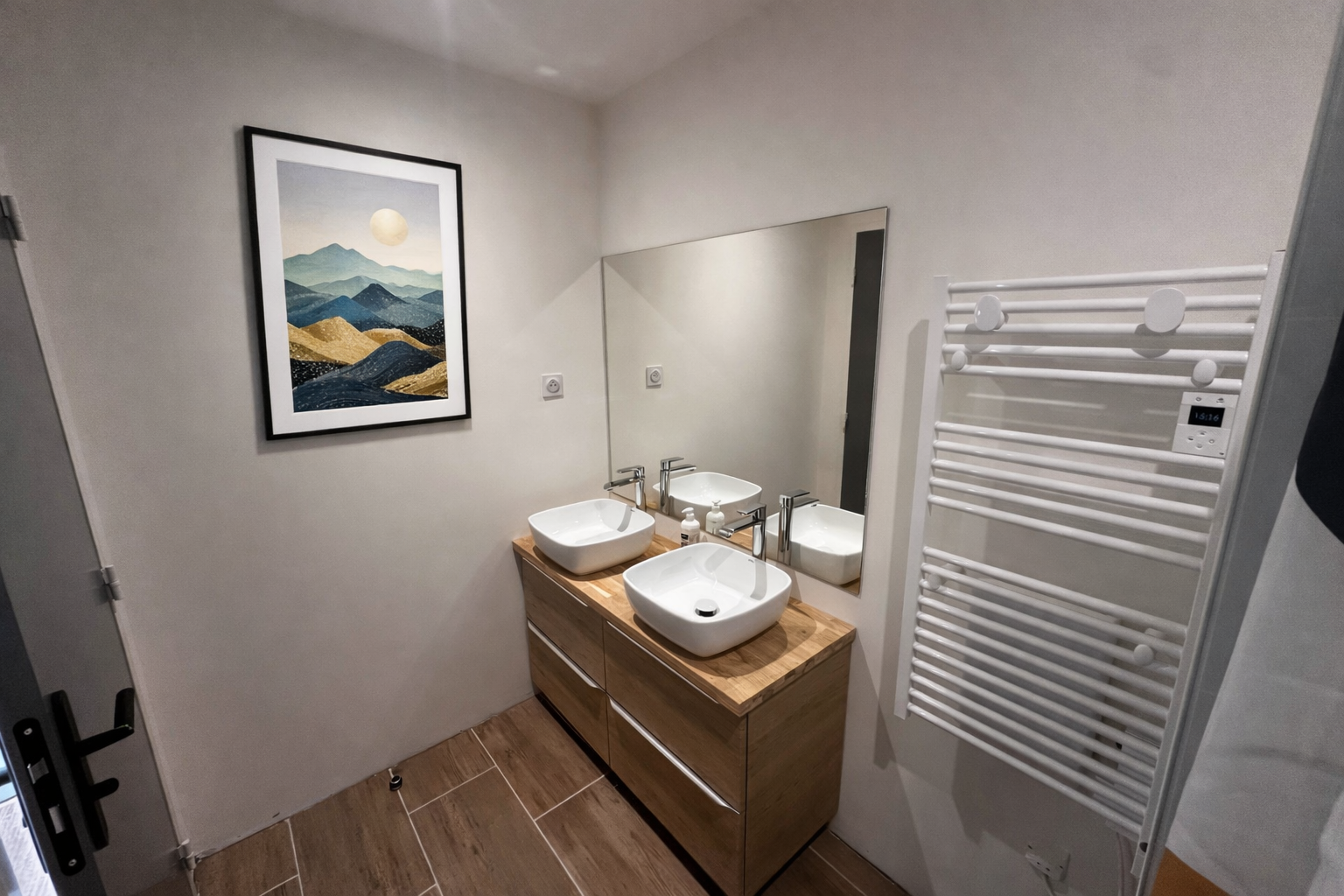 Salle de bain avec deux lavabos en lavabo blanc sur un meuble en bois, un grand miroir, un tableau avec un paysage de montagnes et un soleil, et un radiateur électrique blanc.