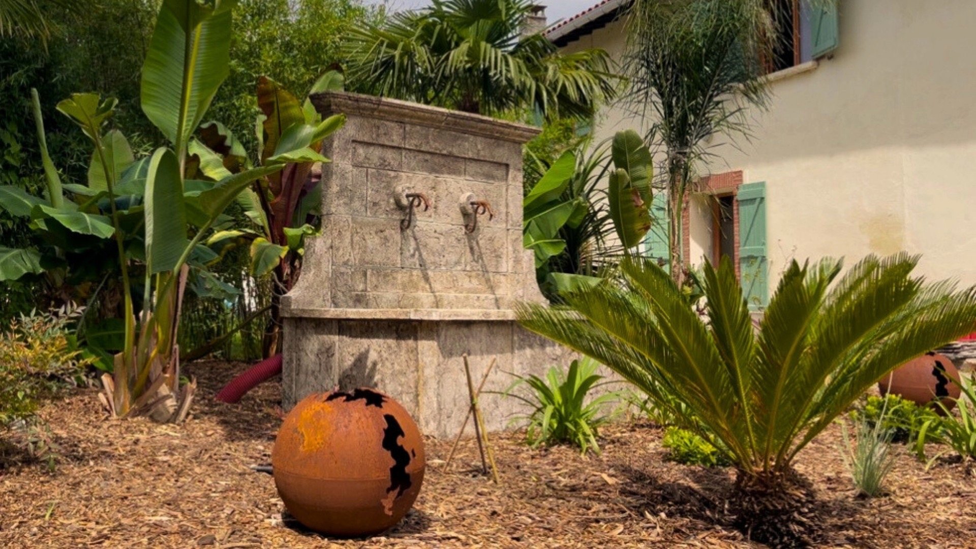 Jardin avec une fontaine en pierre, plantes tropicales, pot en terre cuite rouillée et maison avec volets verts en arrière-plan.