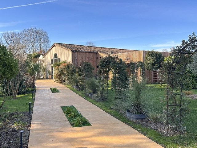 Chemin en béton menant à un bâtiment avec une cour bordée d'arbustes et de plantes, jardin bien entretenu, ciel clair.