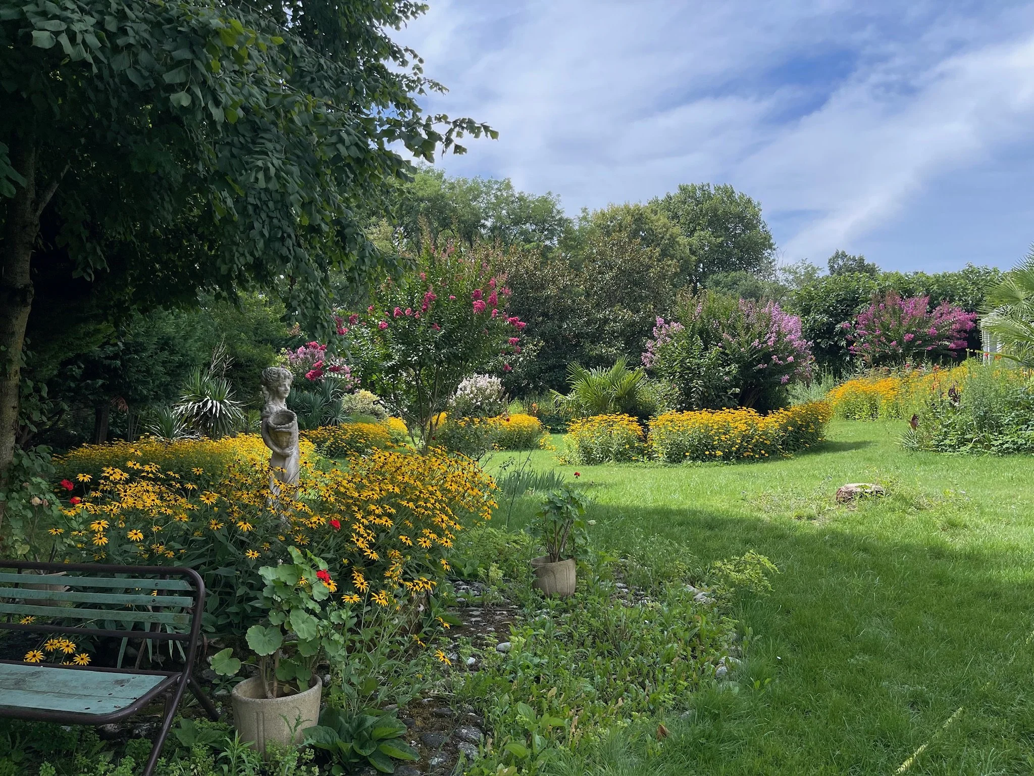 Jardin coloré avec fleurs jaunes, roses, et violettes, statue de femme, banc, et arbres sous un ciel bleu.