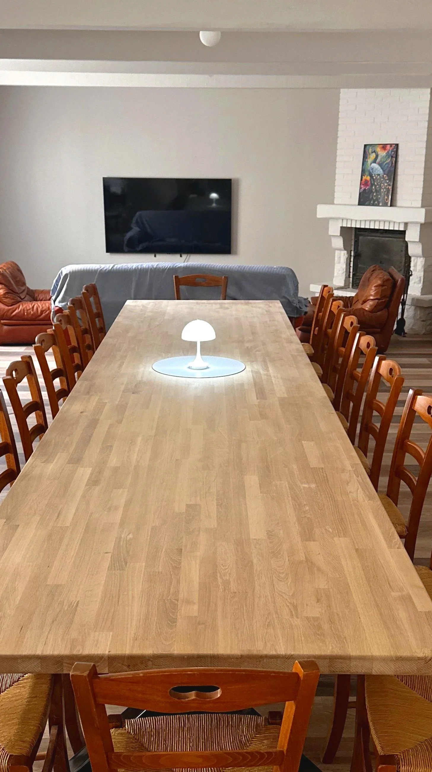 Salle à manger avec une grande table en bois, entourée de chaises en rotin, décorée de bougies et de vases de fleurs, avec une cheminée et un téléviseur à l'arrière, dans une pièce lumineuse avec des fenêtres et des peintures sur les murs.