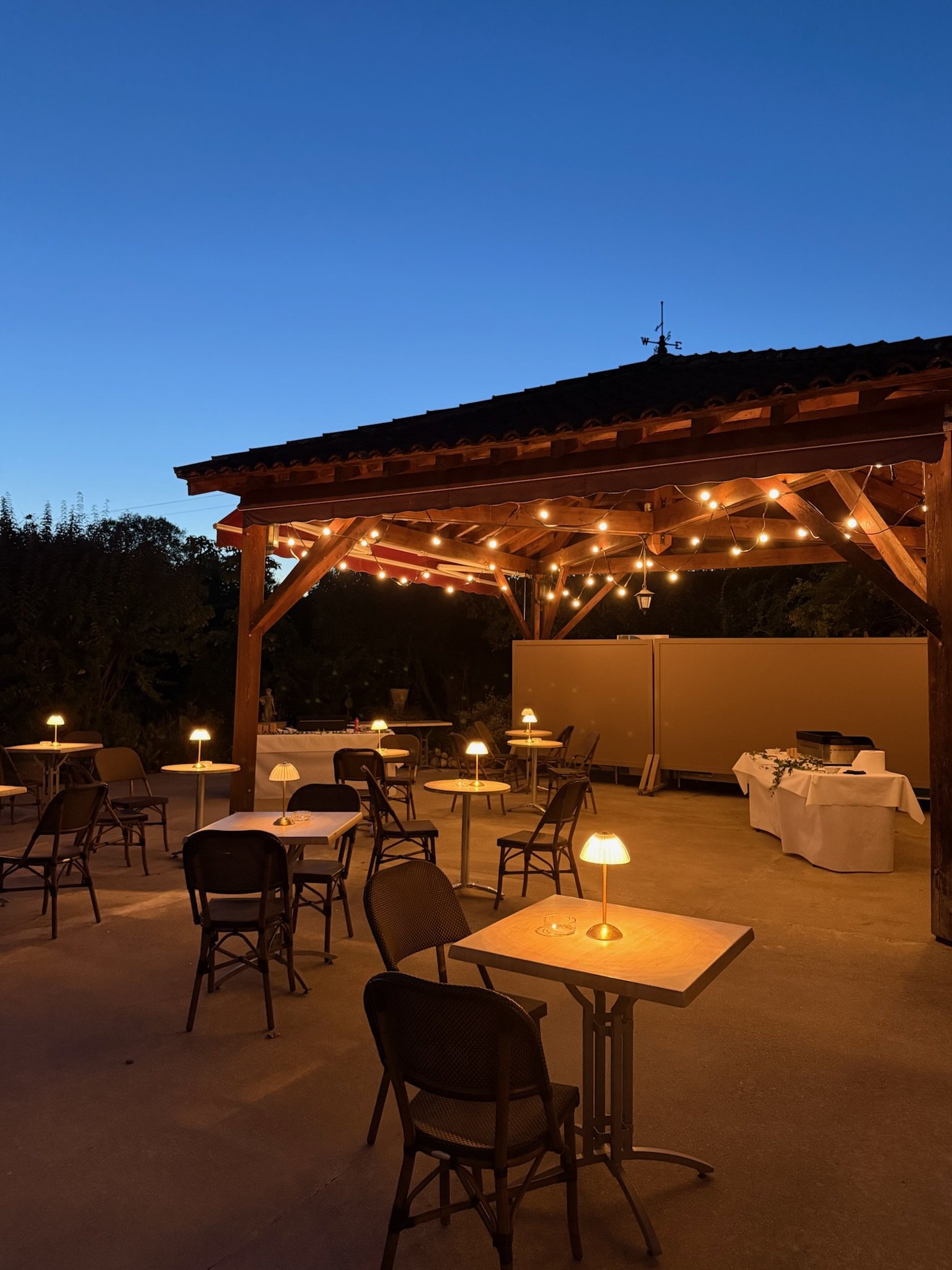 Groupe de personnes dîner en extérieur pendant la soirée, éclairé par des guirlandes lumineuses sous un toit en bois, ciel nocturne bleu en arrière-plan.