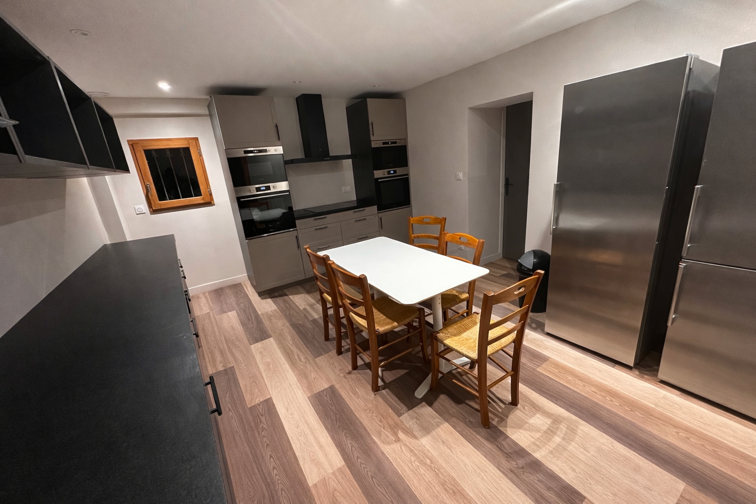 Cuisine avec un plan de travail noir, un réfrigérateur en acier inoxydable, une table blanche avec six chaises en bois, une fenêtre en bois, plusieurs appareils de cuisson encastrés, et un sol en bois.
