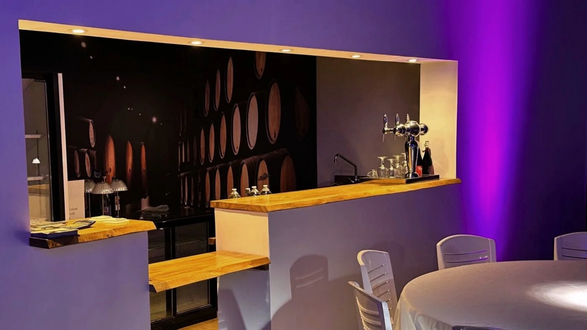 Bar avec comptoir en bois, murs violets, éclairage tamisé, pièces de musique, tables avec chaises, verre à vin, fontaine à bière.