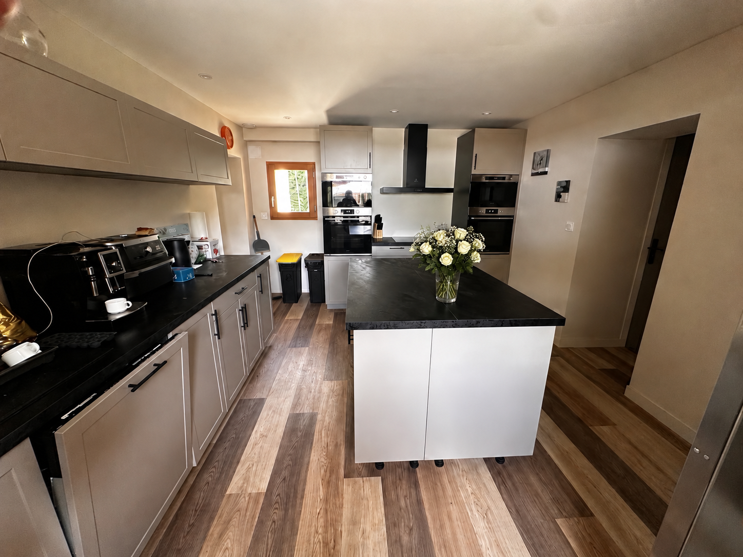 Cuisine avec un plan de travail noir, un réfrigérateur en acier inoxydable, une table blanche avec six chaises en bois, une fenêtre en bois, plusieurs appareils de cuisson encastrés, et un sol en bois.